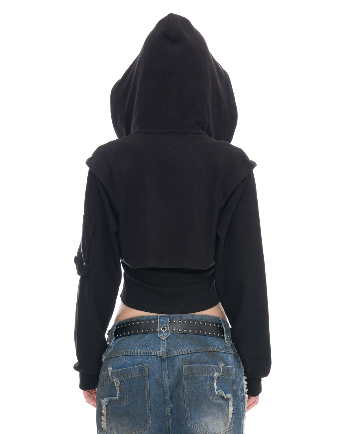 A person shown from the back wearing a black cropped hoodie with long sleeves and a high hood, paired with blue jeans and a black studded belt, against a plain white background.