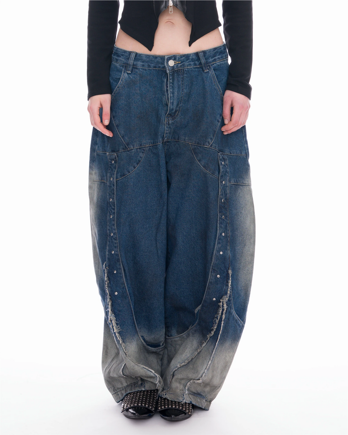 A model is shown from the waist down wearing extremely baggy, wide-leg blue denim jeans with a faded gray gradient at the hems and decorative silver studs running down the front seams.