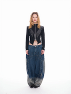 A model stands against a white studio background wearing a black cropped zip-front jacket with dark gray shoulder panels and oversized blue denim pants with gray faded sections and metal rivet details.