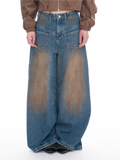 A model is shown from the waist down wearing extremely wide-leg, baggy blue denim jeans with a brown-to-beige gradient wash effect. The model is also wearing a brown cropped jacket and black studded shoes, set against a plain white studio background.
