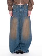A model is shown from the waist down wearing extremely wide-leg, baggy blue denim jeans with a brown-to-beige gradient wash effect. The model is also wearing a brown cropped jacket and black studded shoes, set against a plain white studio background.