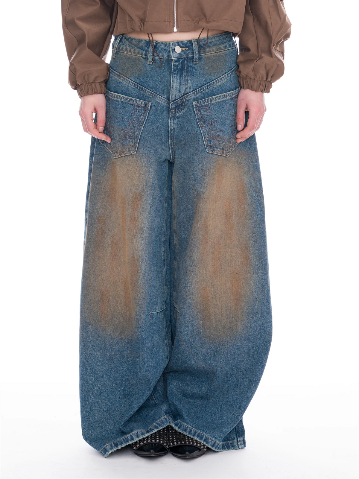 A model is shown from the waist down wearing extremely wide-leg, baggy blue denim jeans with a brown-to-beige gradient wash effect. The model is also wearing a brown cropped jacket and black studded shoes, set against a plain white studio background.