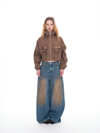 A model stands against a white studio background wearing a cropped brown zip-front jacket with flap pockets and wide-leg faded blue jeans.