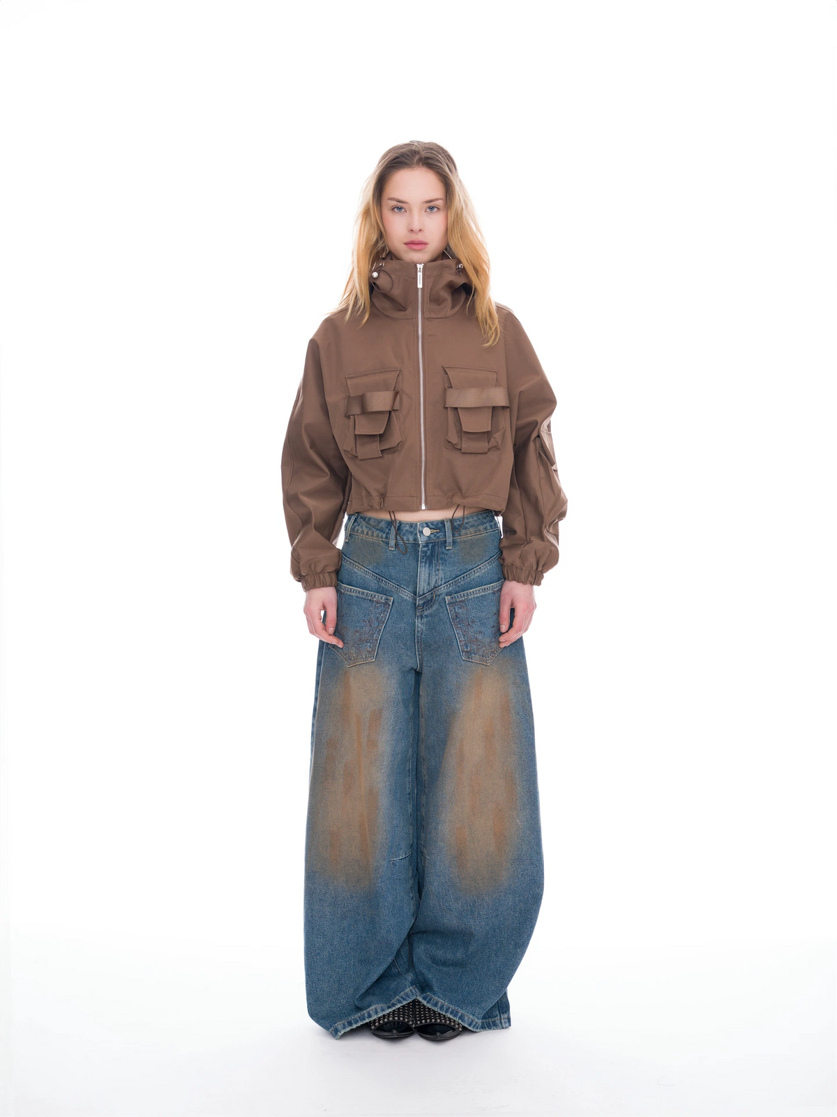 A model stands against a white studio background wearing a cropped brown zip-front jacket with flap pockets and wide-leg faded blue jeans.