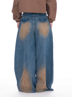 Wide-leg blue denim jeans with a brown tie-dye or bleach-stained pattern, shown from the back on a model wearing a brown cropped jacket against a white background.