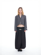 A model stands against a seamless white studio background wearing a dark gray cropped button-front blazer with wide sleeves, black wide-leg trousers, a black belt with a silver buckle, and black pointed flat shoes.
