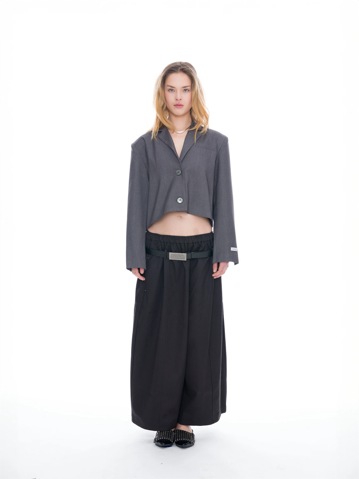 A model stands against a seamless white studio background wearing a dark gray cropped button-front blazer with wide sleeves, black wide-leg trousers, a black belt with a silver buckle, and black pointed flat shoes.