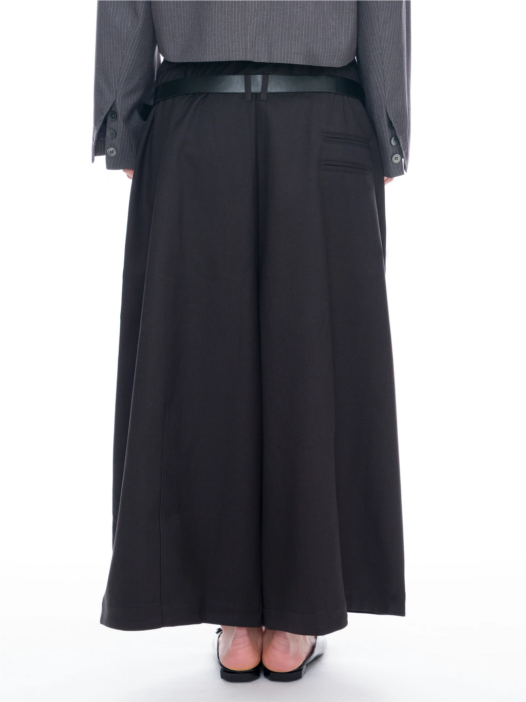 A person shown from the back wearing a long black wide-leg skirt or pant, a dark belt, a gray pinstriped blazer, and black flat shoes against a plain white studio background.
