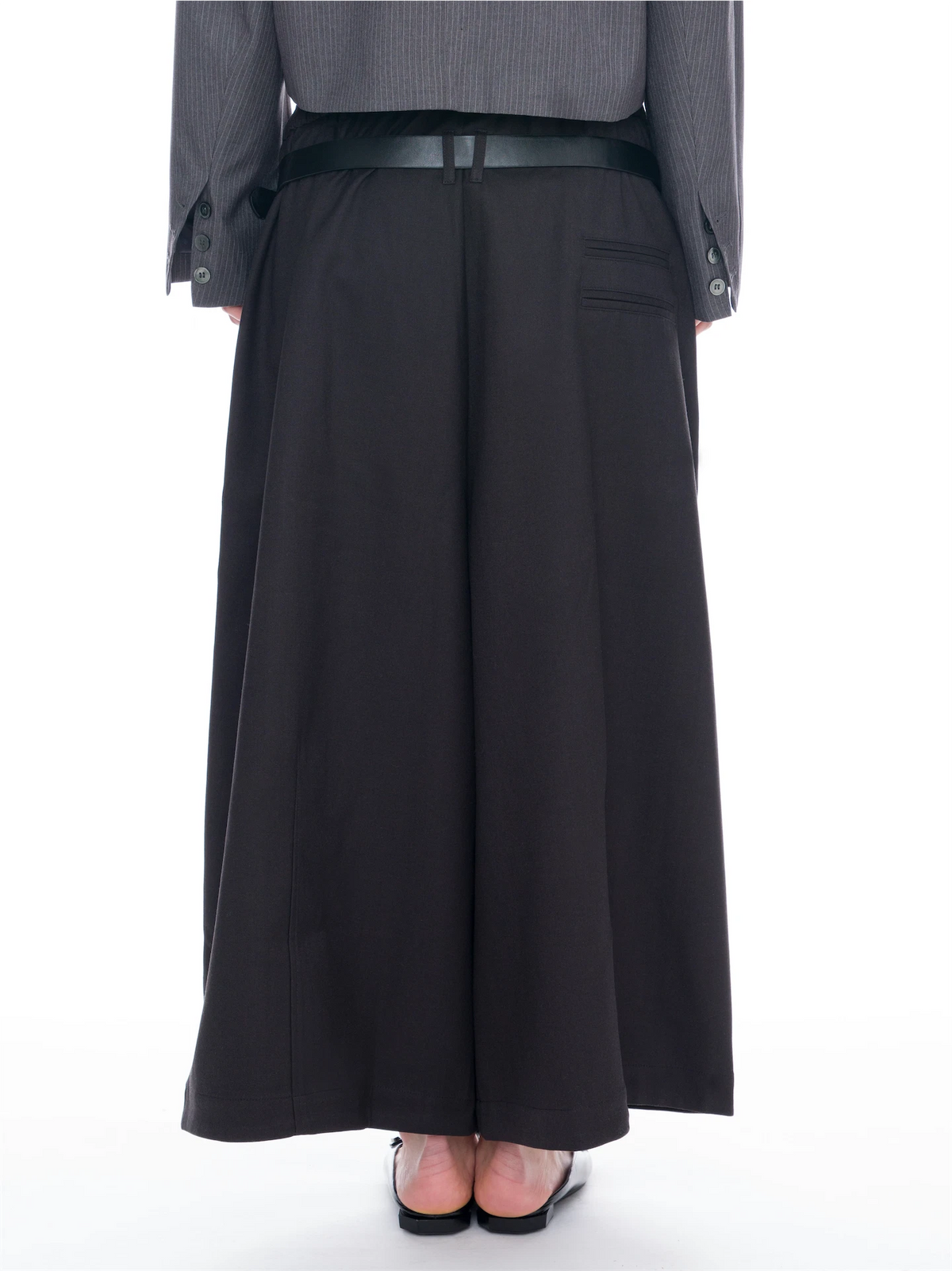 A person shown from the back wearing a long black wide-leg skirt or pant, a dark belt, a gray pinstriped blazer, and black flat shoes against a plain white studio background.