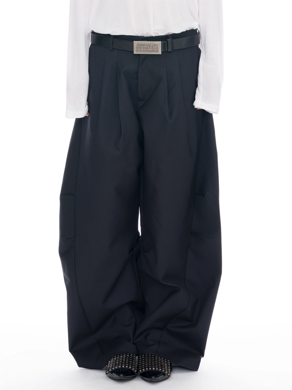 A person wearing very wide-leg black trousers with a black belt, styled with a white long-sleeve top and black studded slide sandals against a plain white background.