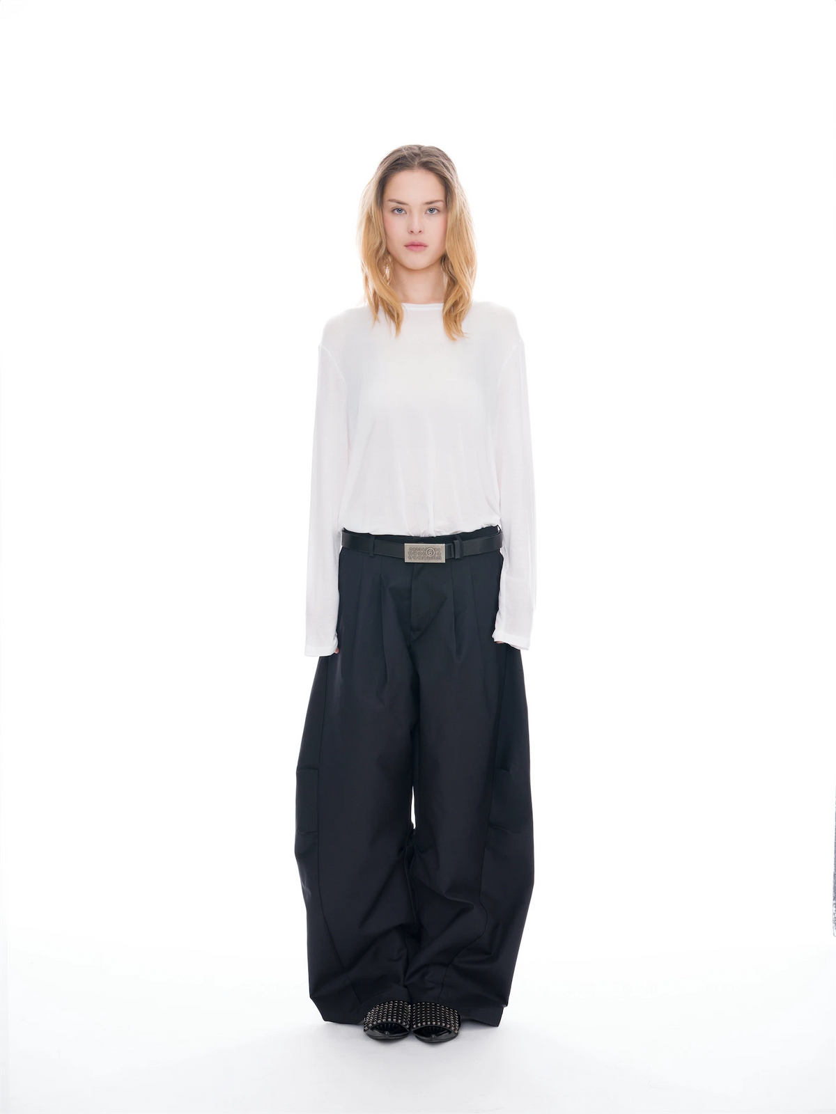 A model stands against a seamless white studio background wearing a white long-sleeve mock-neck top, black wide-leg trousers with a black belt, and black shoes.