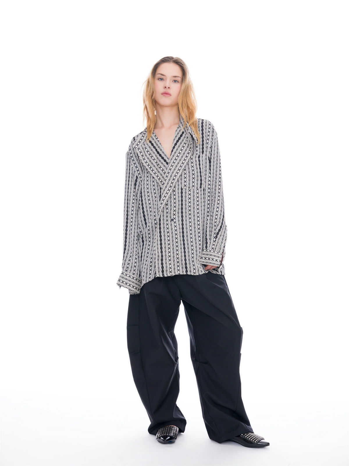 A model stands against a solid white studio background wearing a black and white patterned long-sleeve button-up shirt, loose black wide-leg trousers, and black pointed shoes with silver studs.