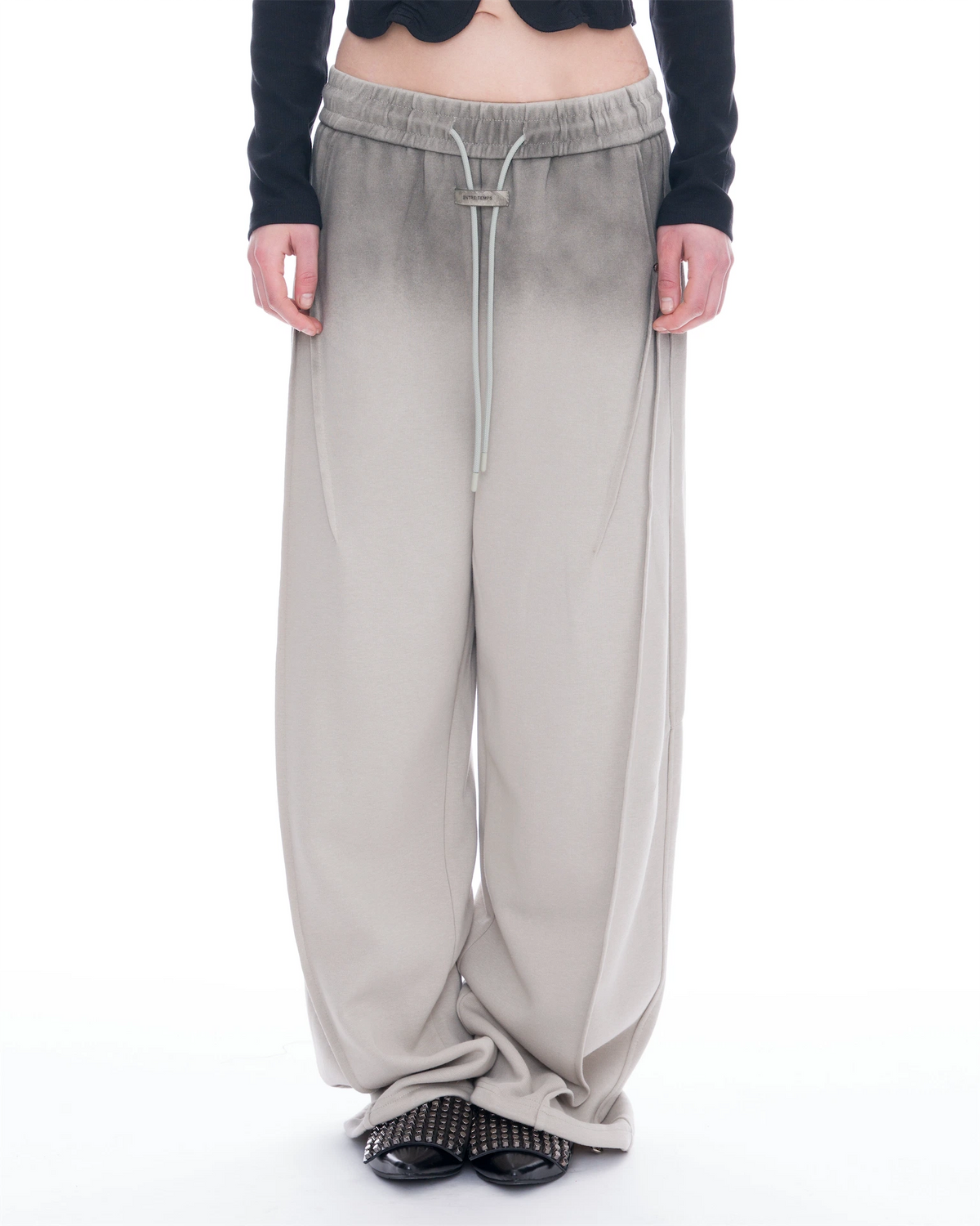 A person wearing wide-leg, floor-length pants with a smooth gradient color transition from black at the top to light gray at the bottom, styled with a black cropped long-sleeve top and black studded flat shoes against a white studio background.