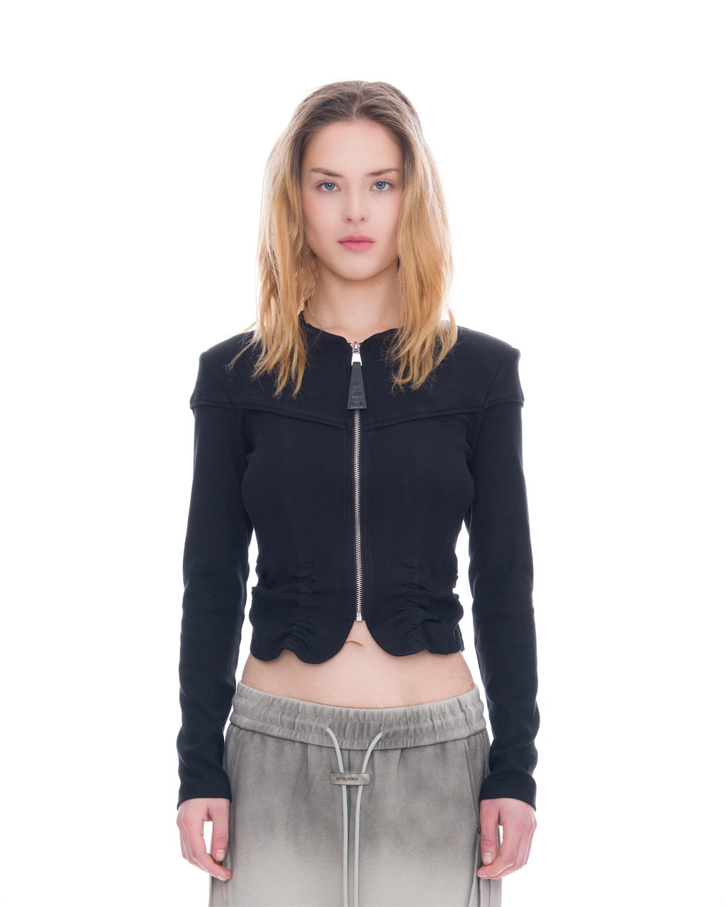 A model wearing a black cropped zip-up jacket with shoulder cutouts and gathered ruffle details at the hem, paired with gray drawstring sweatpants, shot against a solid white background.