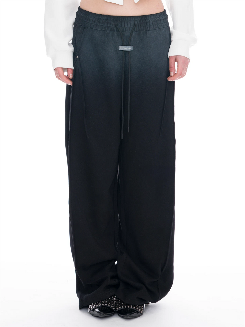 A person wearing wide-leg, floor-length pants with a smooth gradient colour transition from faded black at the top to black at the bottom, styled with a black cropped long-sleeve top and black studded flat shoes against a white studio background.
