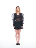 A model stands against a seamless white studio background wearing a black sheer mesh long-sleeve top with dramatic wing-like shoulder panels, paired with black shorts and black studded flat shoes.