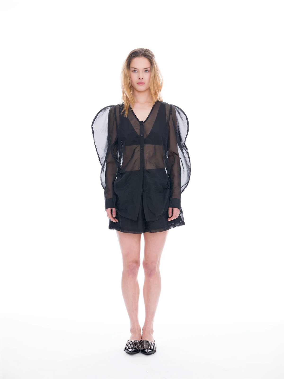 A model stands against a seamless white studio background wearing a black sheer mesh long-sleeve top with dramatic wing-like shoulder panels, paired with black shorts and black studded flat shoes.
