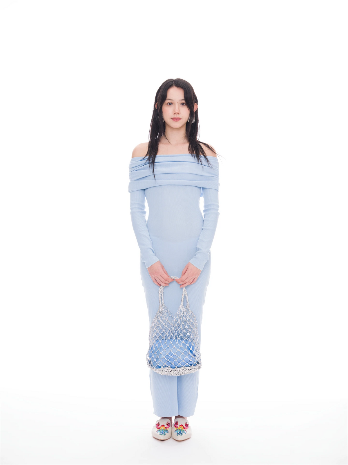 A woman stands against a solid white studio background wearing a light blue off-the-shoulder long-sleeve dress, holding a blue and white net bag, and wearing colorful embroidered shoes.