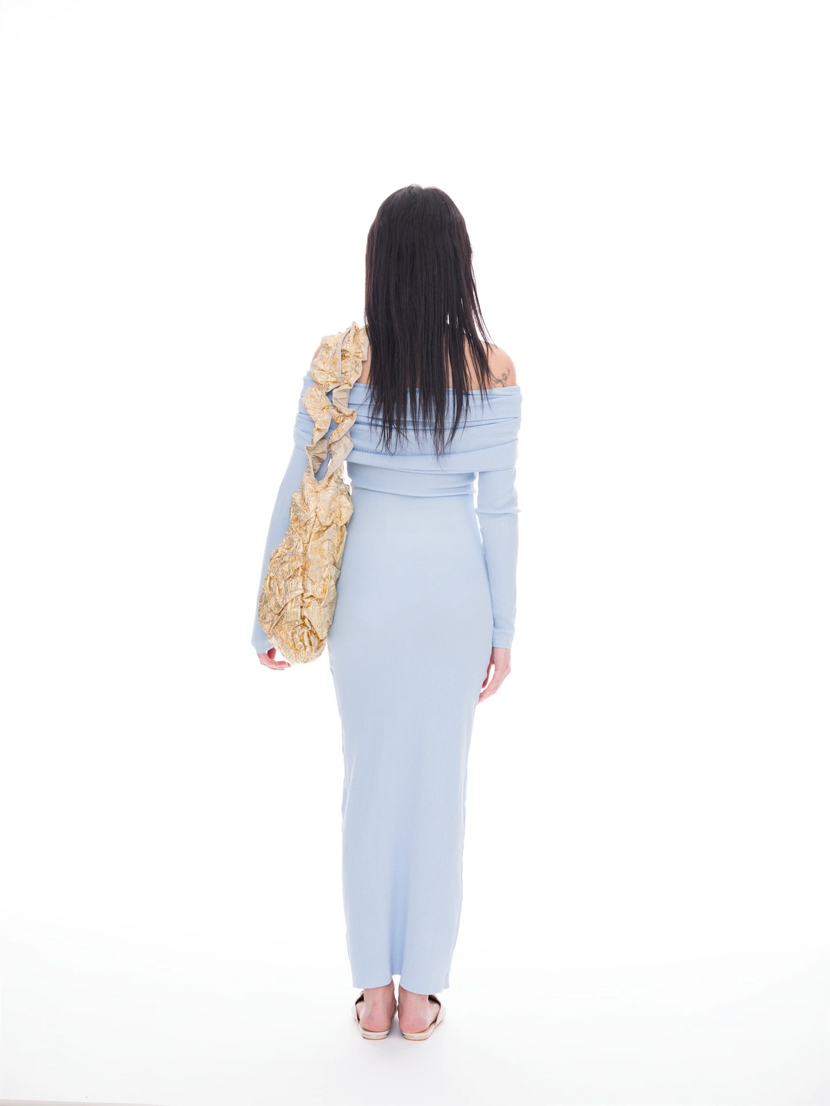 A woman is shown from behind wearing a light blue off-the-shoulder maxi dress and carrying a large beige patterned shoulder bag against a solid white background.