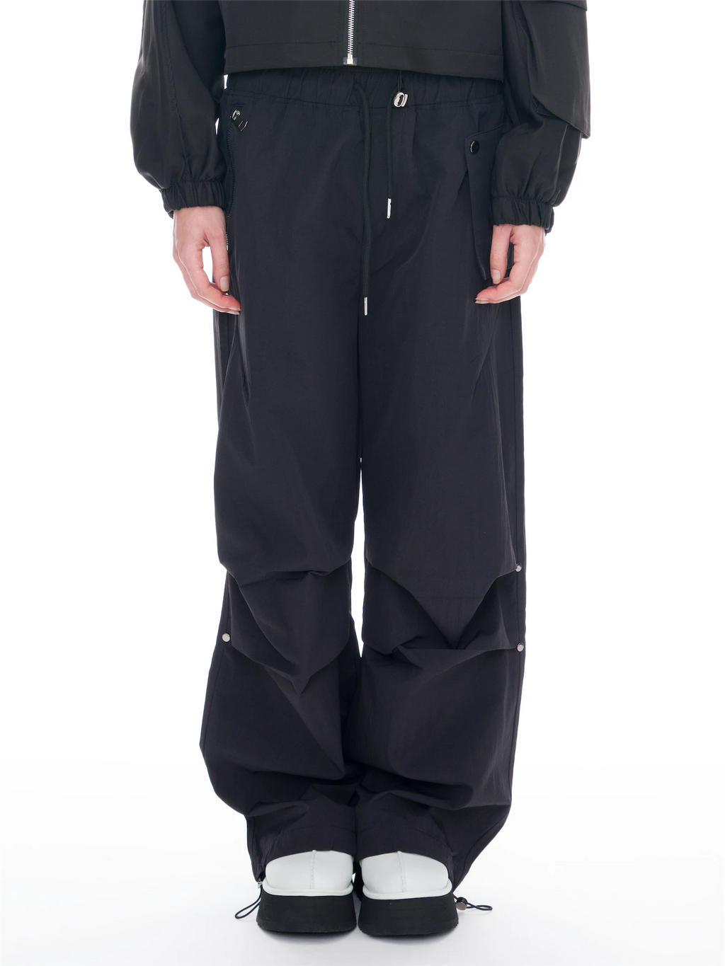 A person wearing black wide-leg cargo pants with a drawstring waist, paired with a black jacket and white-and-black sneakers, photographed against a plain white studio background.
