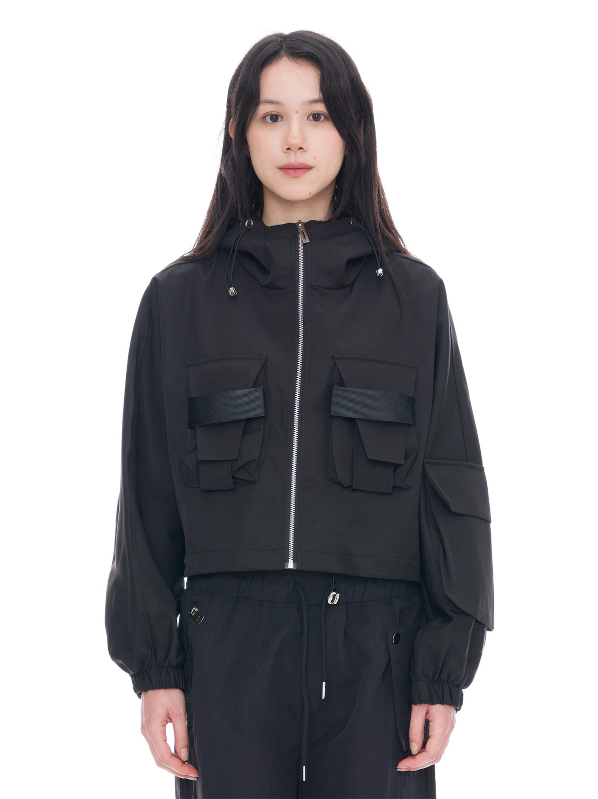 A model wearing a black cropped hooded jacket with a front zipper, two large flap pockets on the chest, and voluminous long sleeves, paired with black pants against a plain white background.