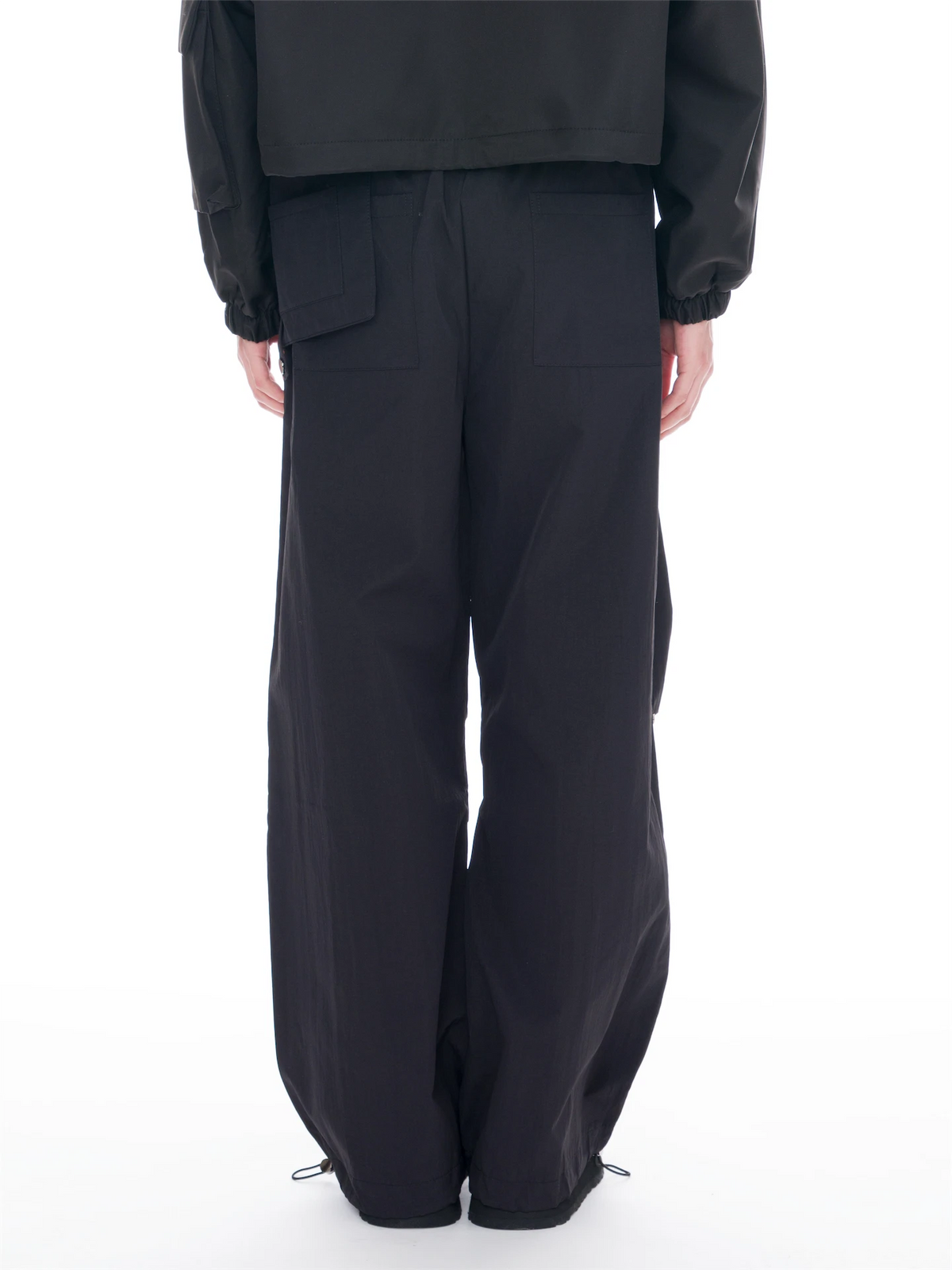 A person shown from the back wearing loose black wide-leg pants with large back patch pockets and a black cropped jacket, photographed against a clean white studio background.