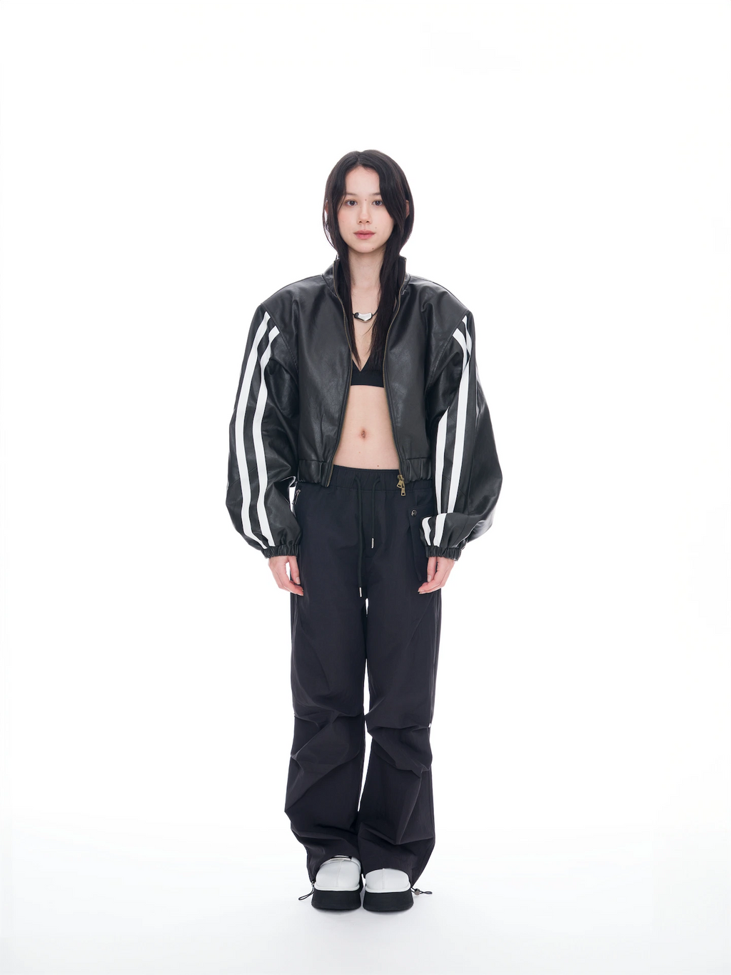 A model stands against a solid white studio background wearing a black leather bomber jacket with white stripes on the sleeves, black pants, and black-and-white sneakers.