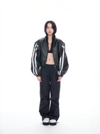 A model stands against a solid white studio background wearing a black leather bomber jacket with white stripes on the sleeves, black pants, and black-and-white sneakers.