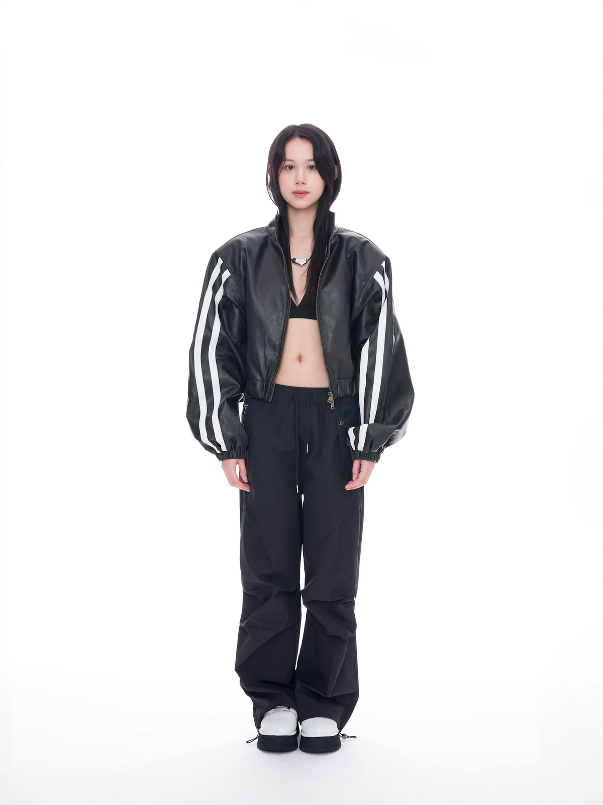 A model stands against a solid white studio background wearing a black leather bomber jacket with white stripes on the sleeves, black pants, and black-and-white sneakers.