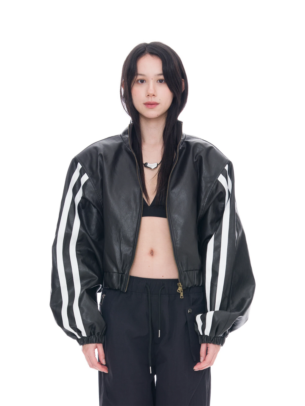 A model wearing a black faux-leather bomber jacket with white stripes on the sleeves, paired with black pants and a black bralette, shot against a solid white background.