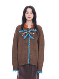 A model wearing a brown knit sweater with a bright blue scalloped trim and a decorative multicolor beaded bow at the chest, paired with a brown and black striped skirt against a solid white background.