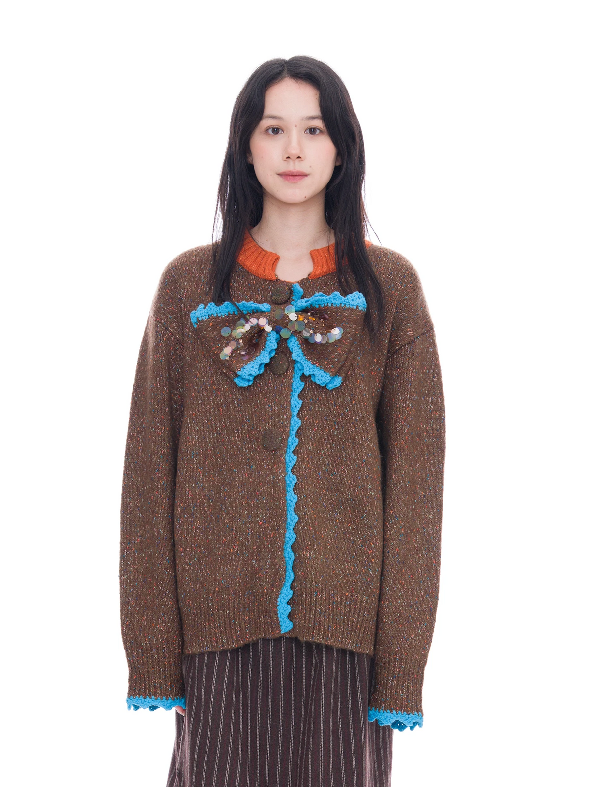 A model wearing a brown knit sweater with a bright blue scalloped trim and a decorative multicolor beaded bow at the chest, paired with a brown and black striped skirt against a solid white background.