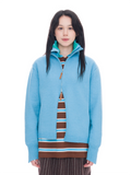 A model wearing a light blue quarter-zip pullover sweater with a teal inner collar, layered over a brown and blue striped knit top and dark pinstriped pants, shot against a solid white background.