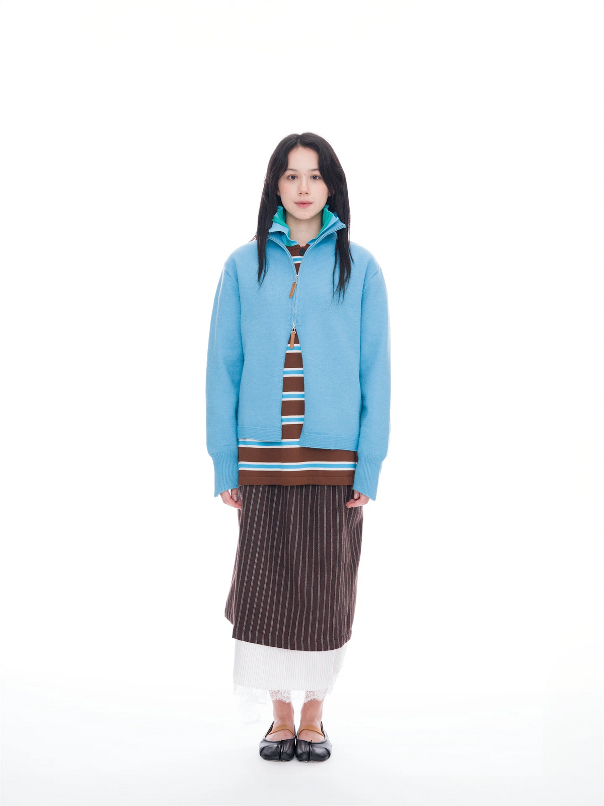 A woman stands against a solid white background wearing a light blue zip-up sweater with brown and white striped accents, a brown pleated skirt, and black shoes.