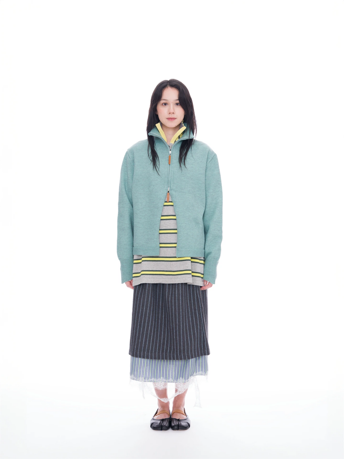 A model stands against a solid white background wearing a light teal-green zip-up sweater layered over a striped knit top, a dark gray pinstriped skirt, and black flat shoes.