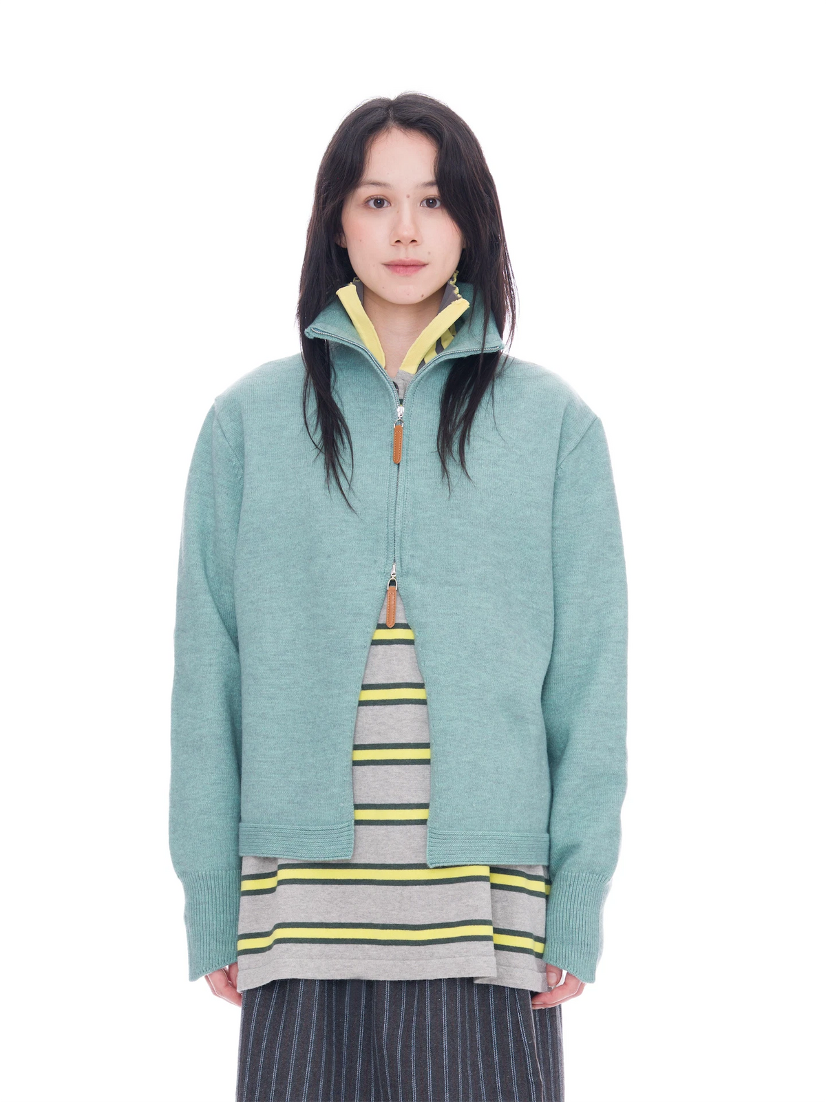 A model wearing a sage green half-zip pullover jacket with a yellow-lined collar, layered over a gray striped shirt and dark pinstriped pants, shot against a solid white background.
