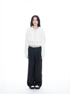 A model stands against a solid white studio background wearing a white cropped zip-up hoodie, black wide-leg pants, and black-and-white slip-on shoes.