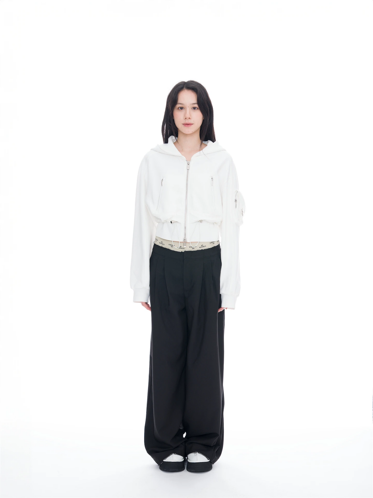 A model stands against a solid white studio background wearing a white cropped zip-up hoodie, black wide-leg pants, and black-and-white slip-on shoes.