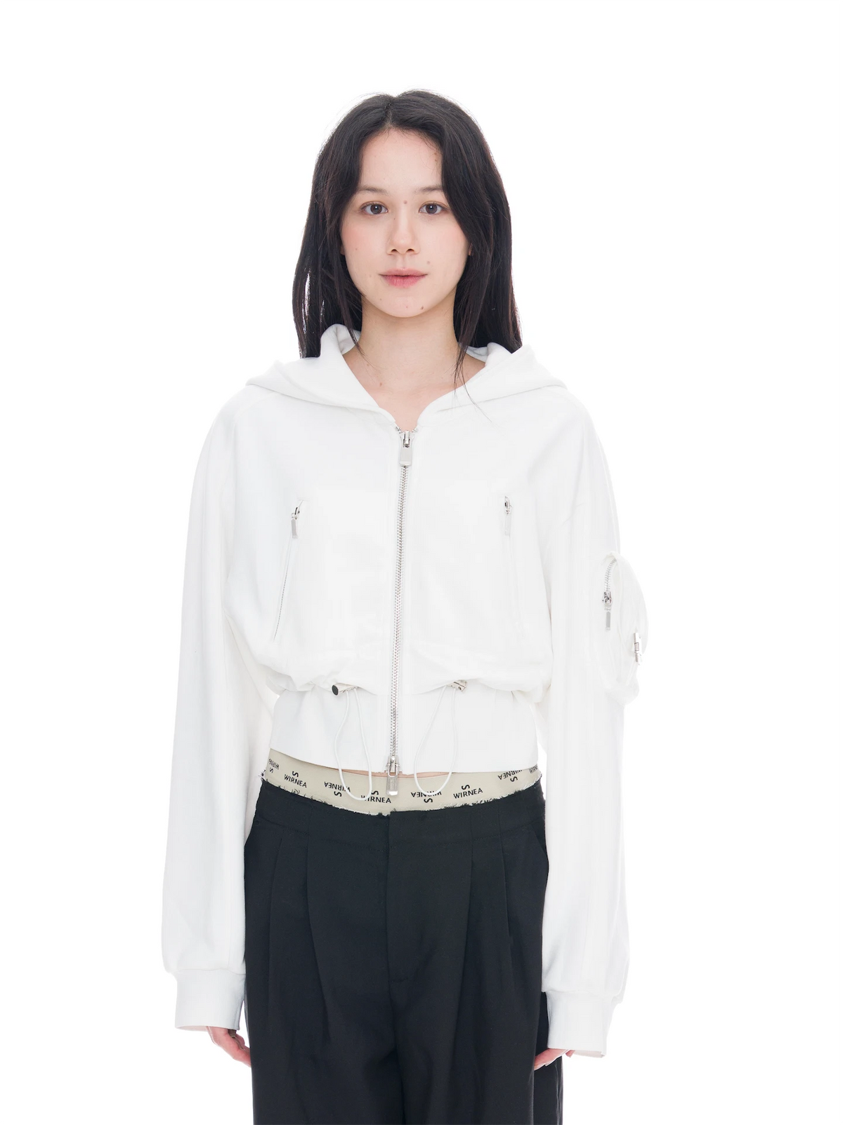 A model wearing a white cropped zip-up hoodie with a hood and front pockets, paired with black trousers, against a solid white background.