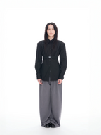 A model stands against a solid white studio background wearing a black structured long-sleeve shirt with a belt detail and wide-leg gray trousers.