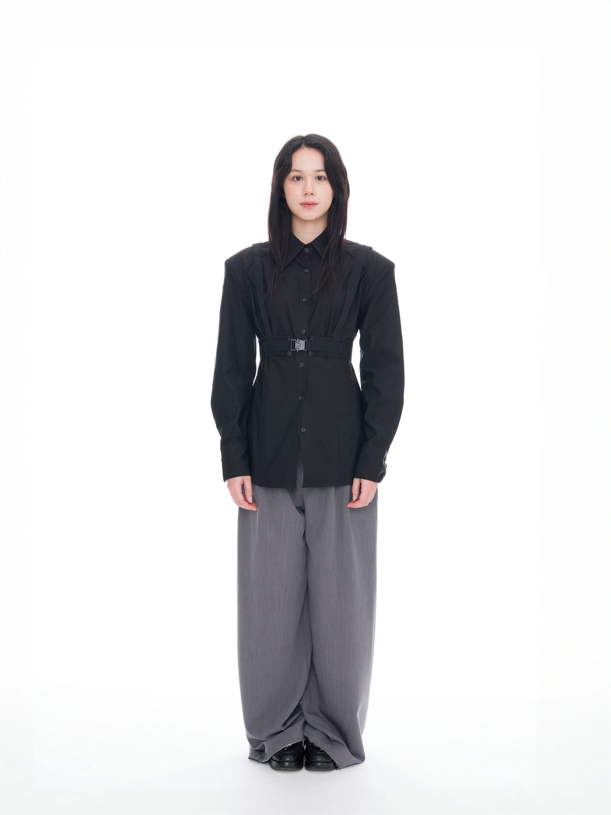 A model stands against a solid white studio background wearing a black structured long-sleeve shirt with a belt detail and wide-leg gray trousers.