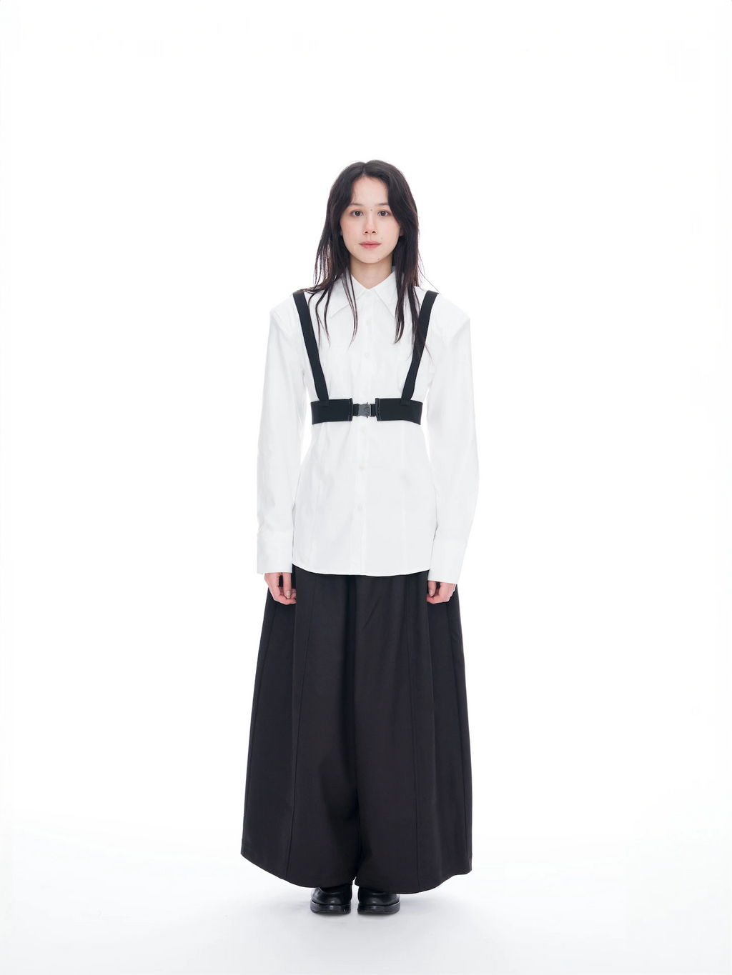 A model stands against a seamless white studio background wearing a white long-sleeve button-up shirt with black suspenders and a black belt detail, paired with wide-leg black trousers.