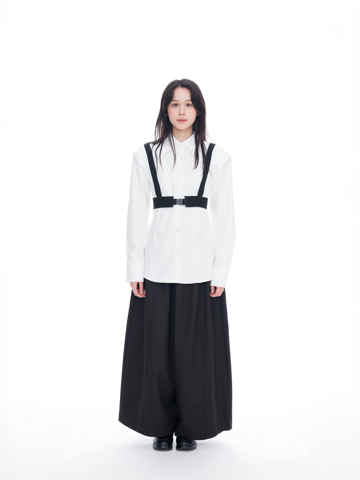 A model stands against a seamless white studio background wearing a white long-sleeve button-up shirt with black suspenders and a black belt detail, paired with wide-leg black trousers.