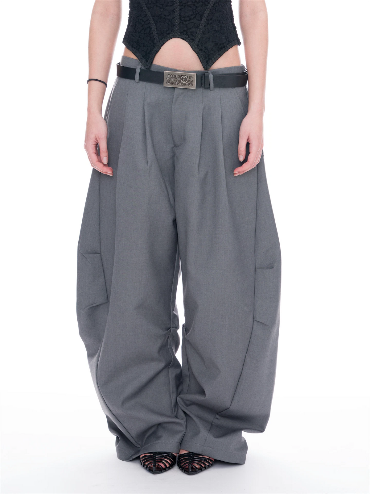 A model is shown from the waist down wearing very wide-leg, baggy gray trousers with a black belt and a silver rectangular buckle. The background is a clean white studio backdrop.