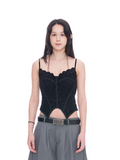 A woman modeling a black lace corset-style camisole top with thin straps and a sheer mesh panel, paired with gray trousers and a black belt, against a plain white background.