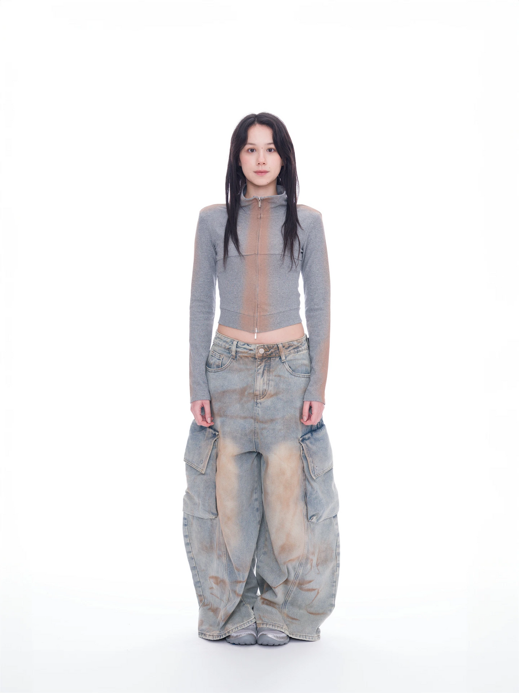 A model stands against a seamless white studio background wearing a gray mock-neck long-sleeve crop top with a sheer mesh front panel, paired with extremely baggy light-wash denim cargo pants featuring a brown vertical stripe down the front of each leg.