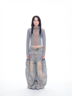 A model stands against a seamless white studio background wearing a gray mock-neck long-sleeve crop top with a sheer mesh front panel, paired with extremely baggy light-wash denim cargo pants featuring a brown vertical stripe down the front of each leg.