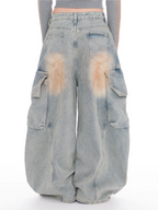A person shown from the back wearing very baggy light-wash denim cargo pants with large side pockets and faded tan staining on the back pockets, against a plain white studio background.