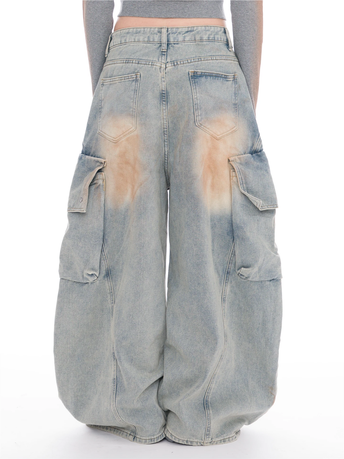 A person shown from the back wearing very baggy light-wash denim cargo pants with large side pockets and faded tan staining on the back pockets, against a plain white studio background.