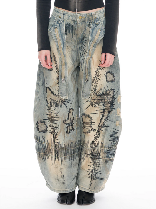 A person wearing extremely baggy distressed denim pants with abstract black painted markings, a black long-sleeve top, and black shoes, photographed against a plain white studio background.